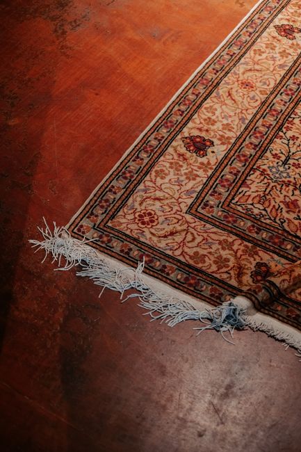 A close-up view of a modern cordless vacuum cleaner with a black and red design, positioned on a detailed, patterned rug with shades of orange, blue, and beige, in a well-lit room. The vacuum head is 