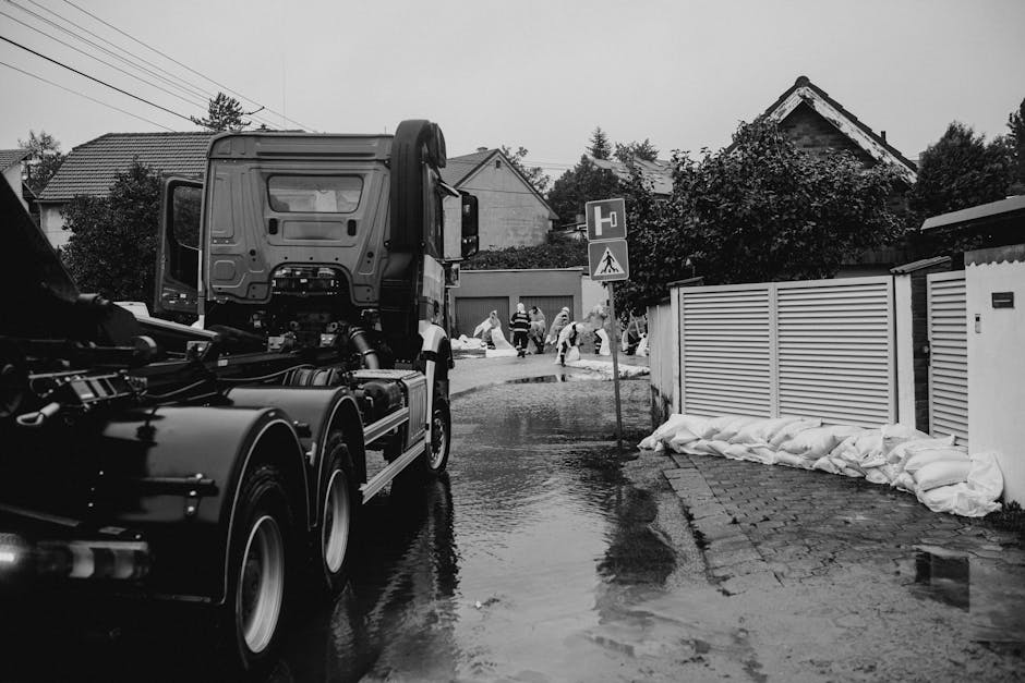 A residential street scene in black and white showing a large truck parked on the wet pavement during a flood response. In the background, several workers are seen wearing protective gear and engaging